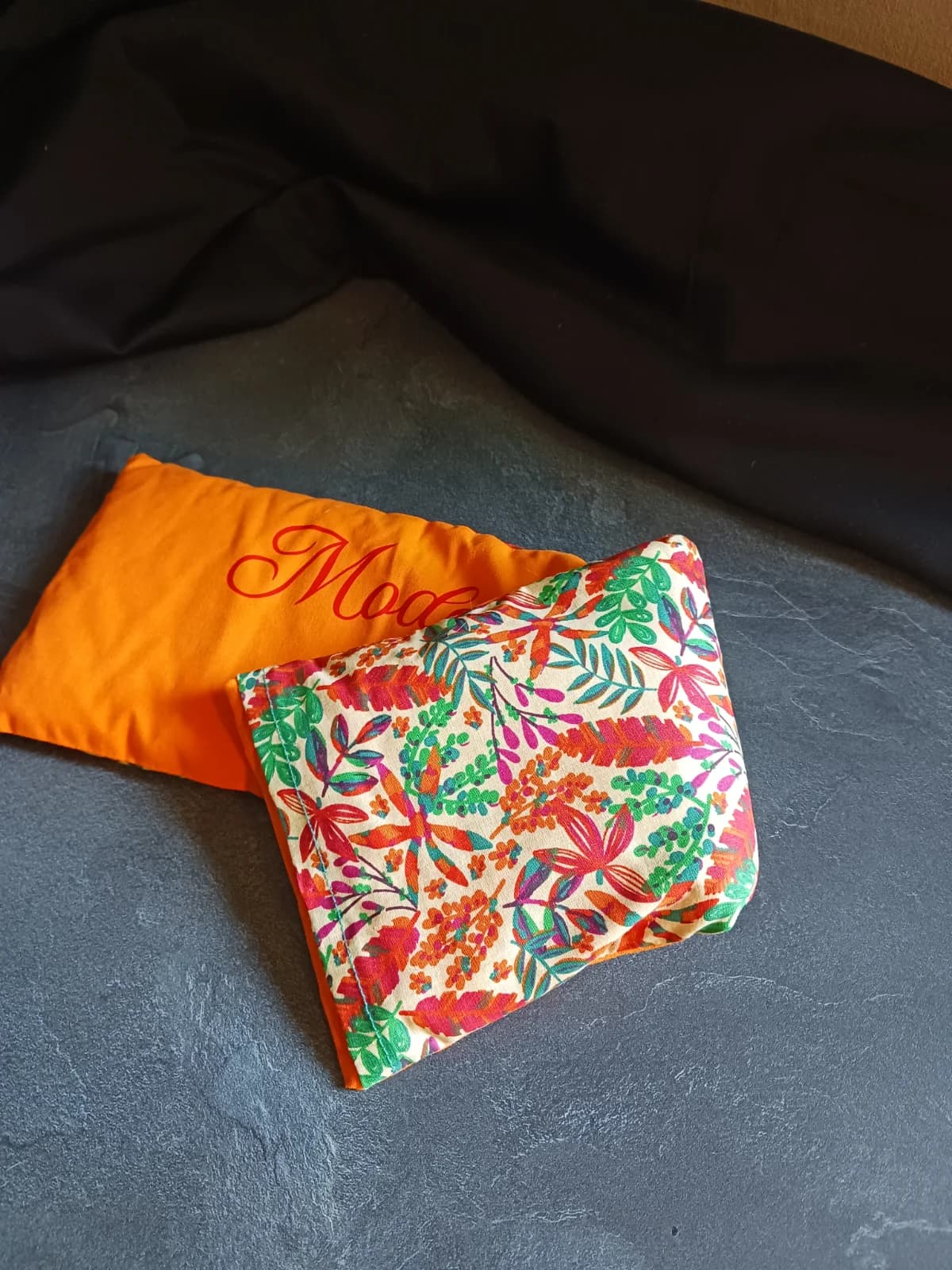 Bouillotte pliée laissant voir la housse orange et le coussin à motif végétal multicolore.